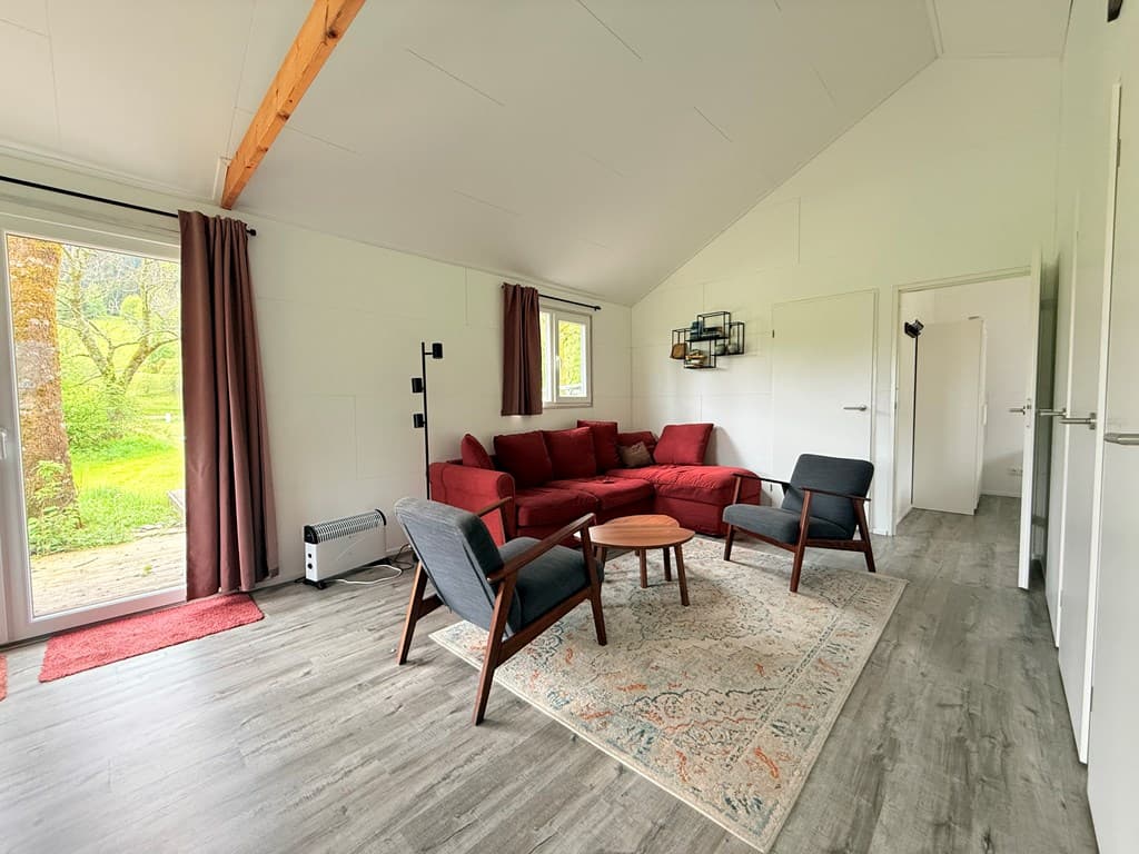 Woonkamer met een rood bankstel, twee fauteuils en toegang tot een tuin via een terrasdeur.