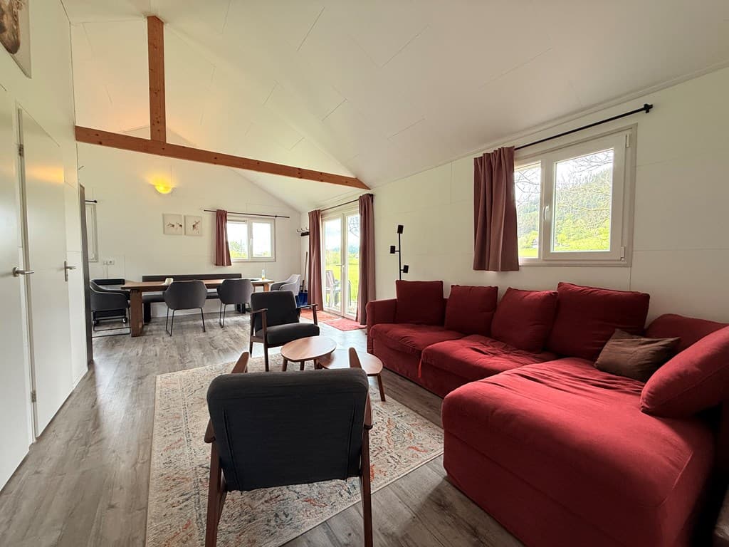 Ruime woonkamer met een rode bank, eettafel en stoelen bij een raam met uitzicht op de natuur.