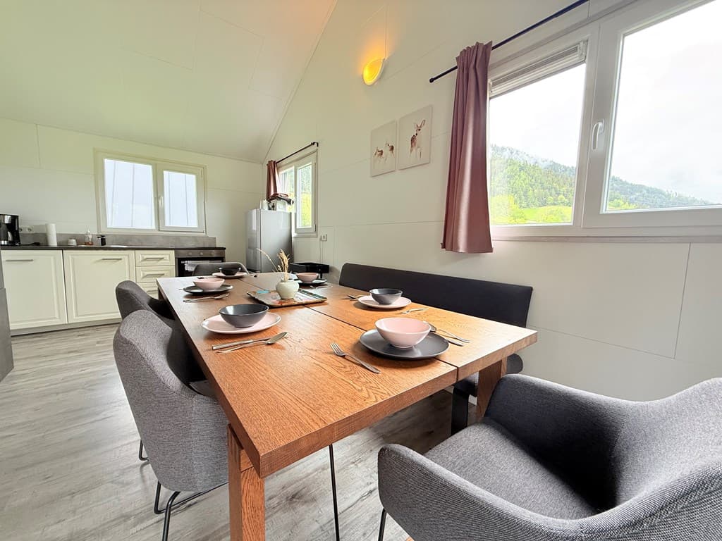 Eetkamer met een grote houten tafel, zes stoelen en uitzicht op groene heuvels door de ramen.