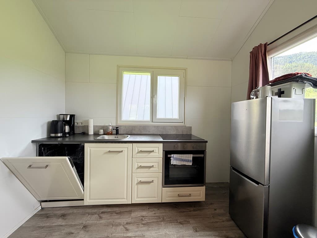 Moderne, lichte keuken met witte kasten, zwarte werkblad en grote koelkast naast een raam met uitzicht op groen.