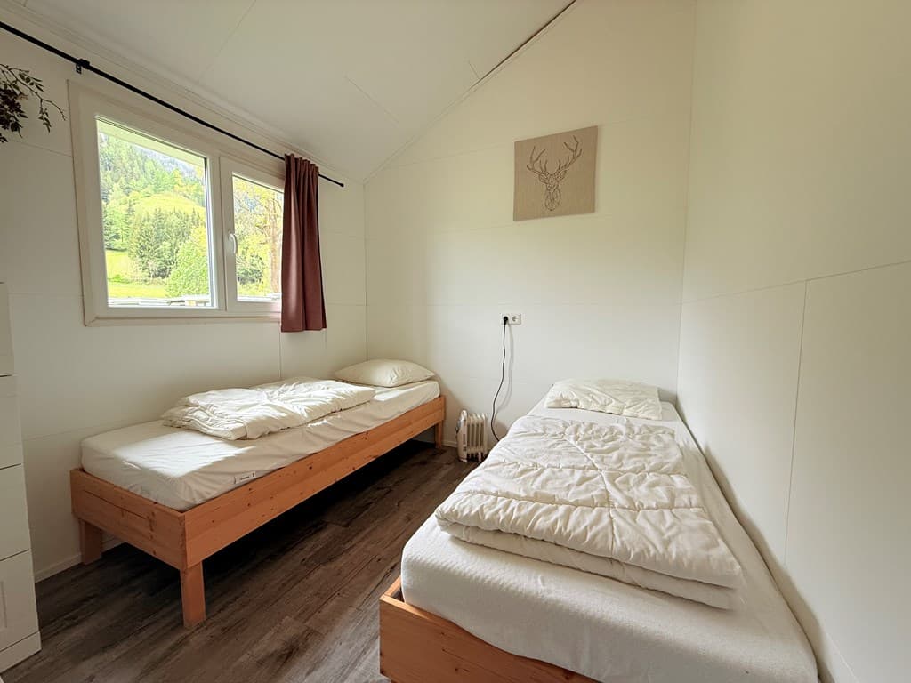 Twee eenvoudige eenpersoonsbedden in een lichte kamer met houten vloeren en uitzicht op groene heuvels buiten.