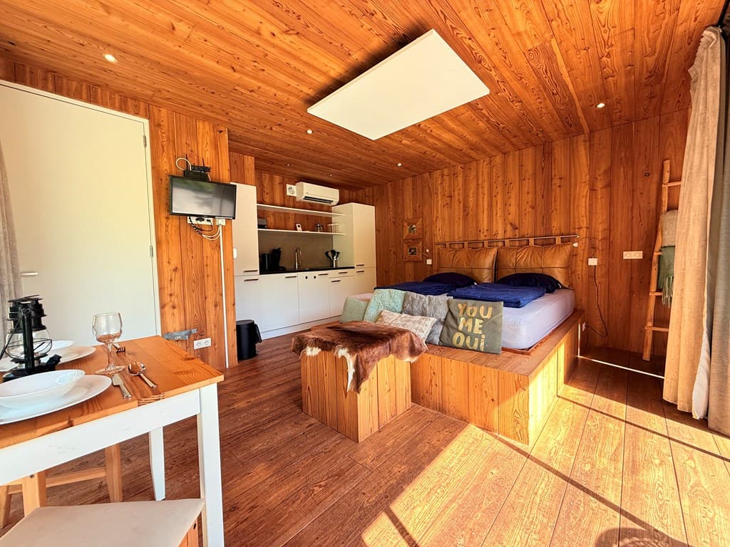 Houten interieur van een vakantiewoning met een bed, eettafel en zithoek, grote ramen zorgen voor veel lichtinval