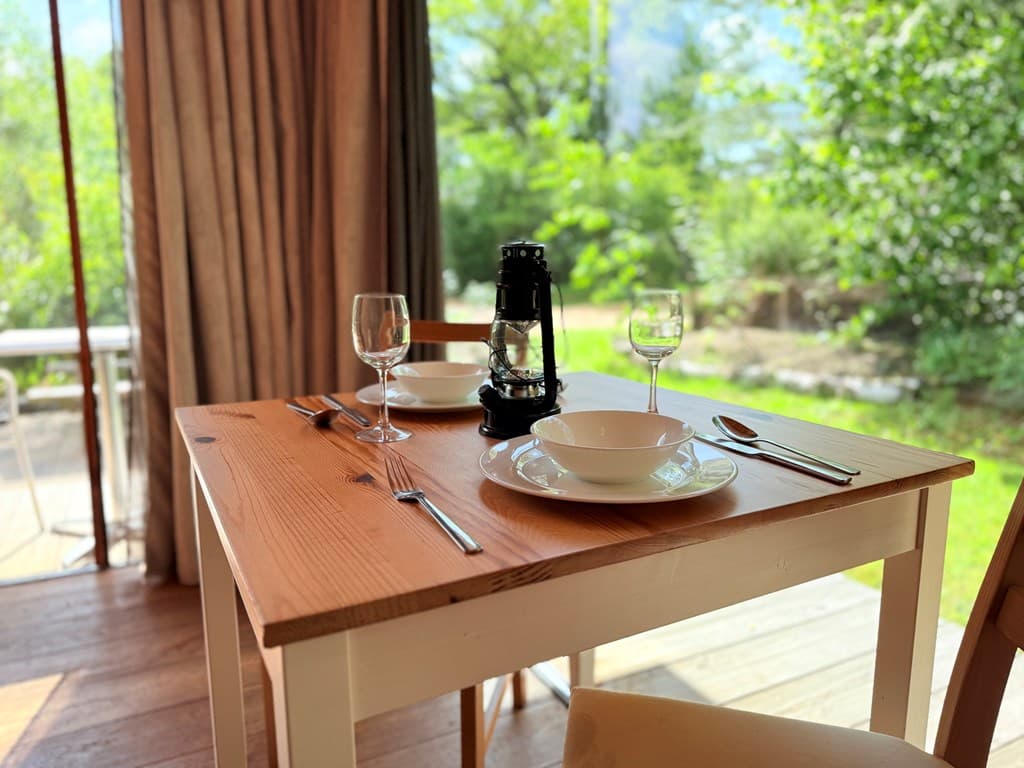 Eetkamertafel met borden, glazen en een lampje, uitzicht op groene omgeving door grote ramen