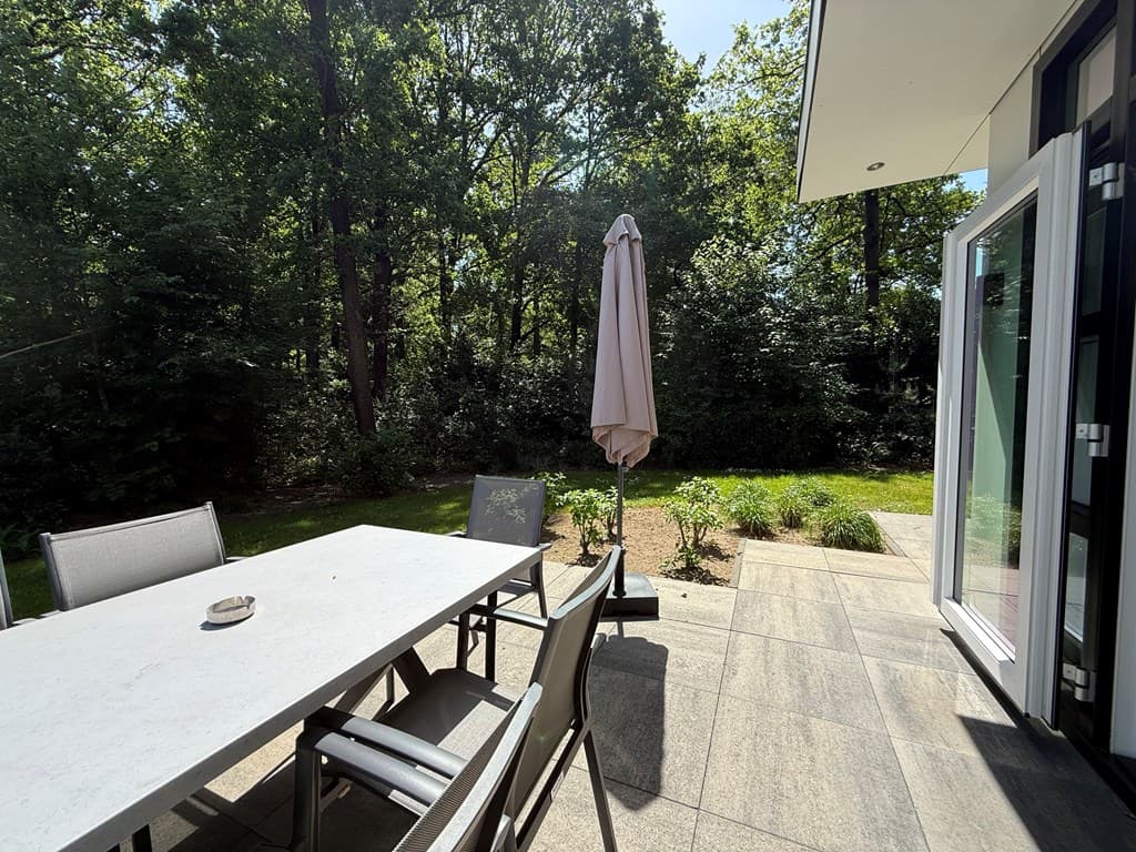 Terras met een tafel en stoelen, een parasol en uitzicht op een groene, bosachtige tuin.