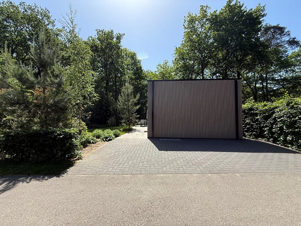 Moderne vakantiewoning met houten gevel, omringd door groen en een betegelde oprit, gelegen in een bosrijke omgeving