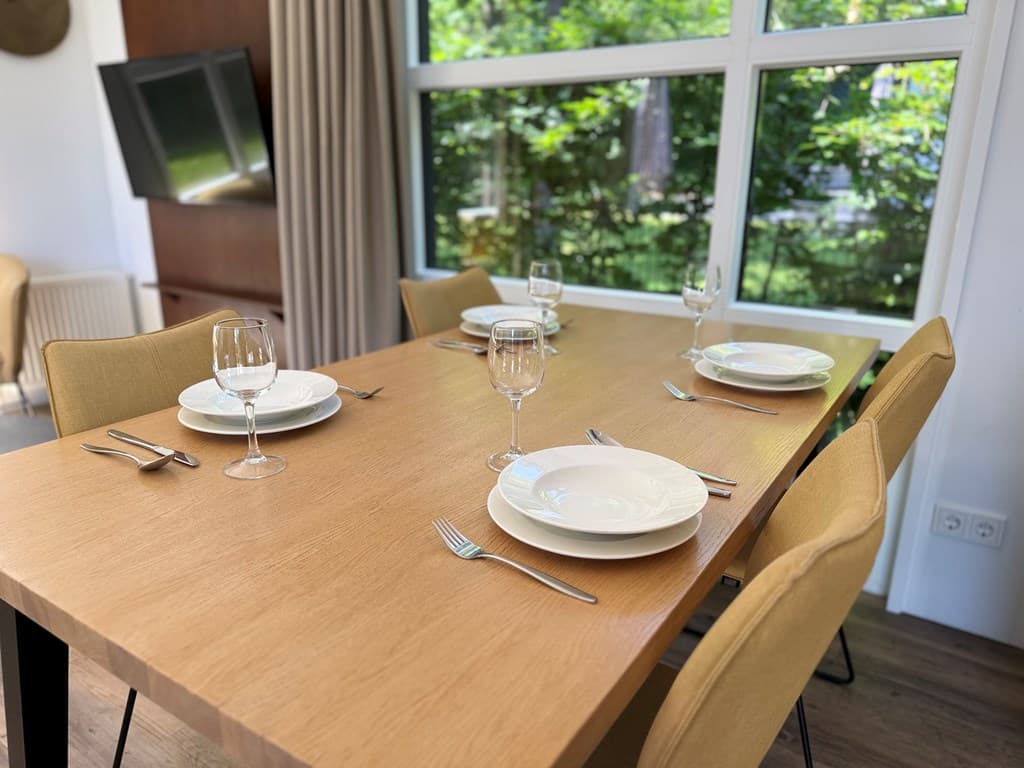 Eettafel met vier borden, glazen en bestek, omgeven door grote ramen met uitzicht op groen.