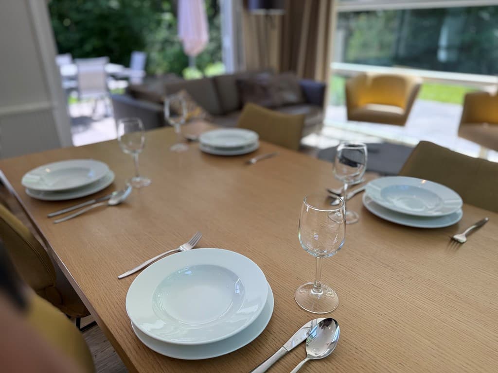 Eettafel met servies en glazen, bank en stoelen zichtbaar op de achtergrond bij een grote raam met uitzicht op de tuin