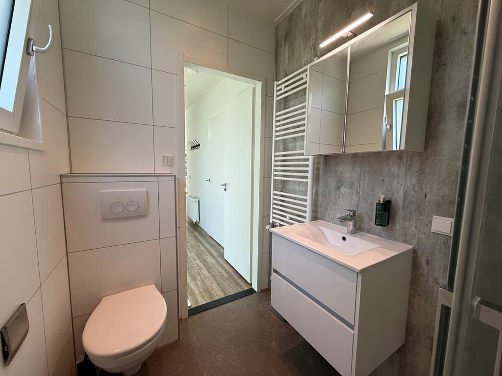 Modern badkamer met wastafel, toilet en spiegels, wand met tegels en toegang tot een andere kamer.