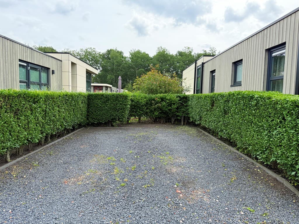 Tussen twee vakantiewoningen staat een betegeld terrein omringd door een laurierhaag en bomen op de achtergrond.