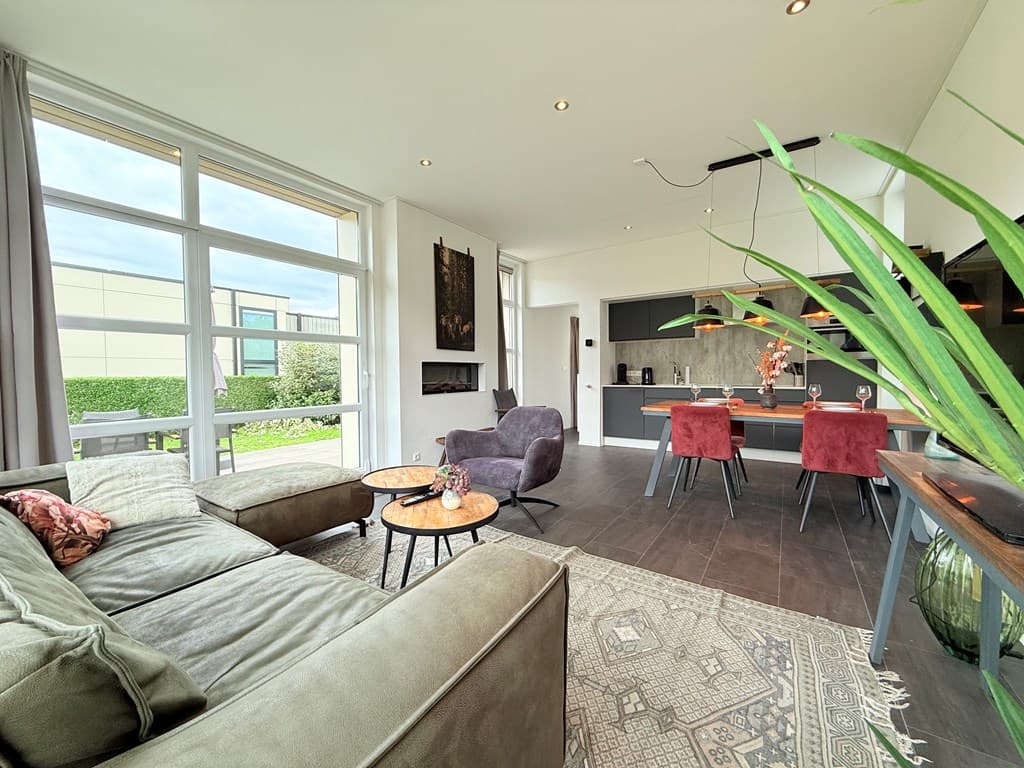 Moderne woonkamer met grote ramen, zithoek, open keuken en eettafel met stoelen, groen uitzicht door de ramen.
