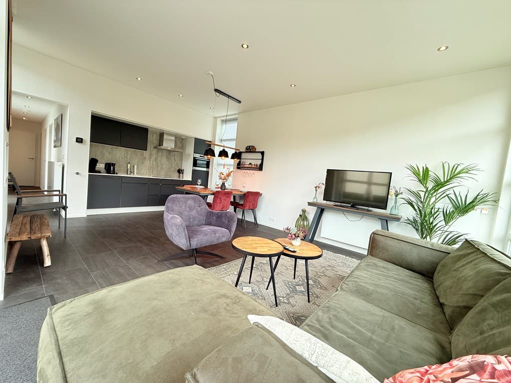 Moderne woonkamer met bank, salontafels, tv en eettafel met stoelen, lichte kleuren en open keukengedeelte zichtbaar
