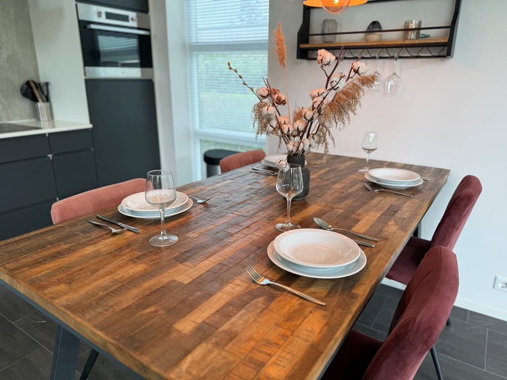 Eetkamer met een houten tafel, vier stoelen, borden en glazen, en een vaas met droogbloemen op de tafel.