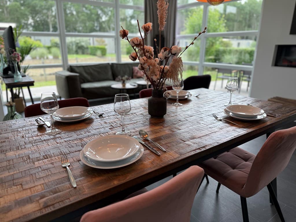 Eetkamer met een lange houten tafel, vier borden en glazen, zicht op een gezellige zithoek en grote ramen naar buiten.