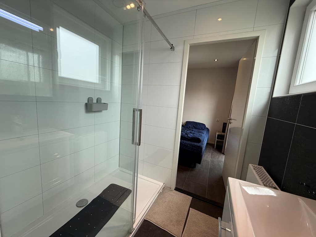 Moderne badkamer met inloopdouche, wastafel en toegang naar een slaapkamer met bed en stoel.