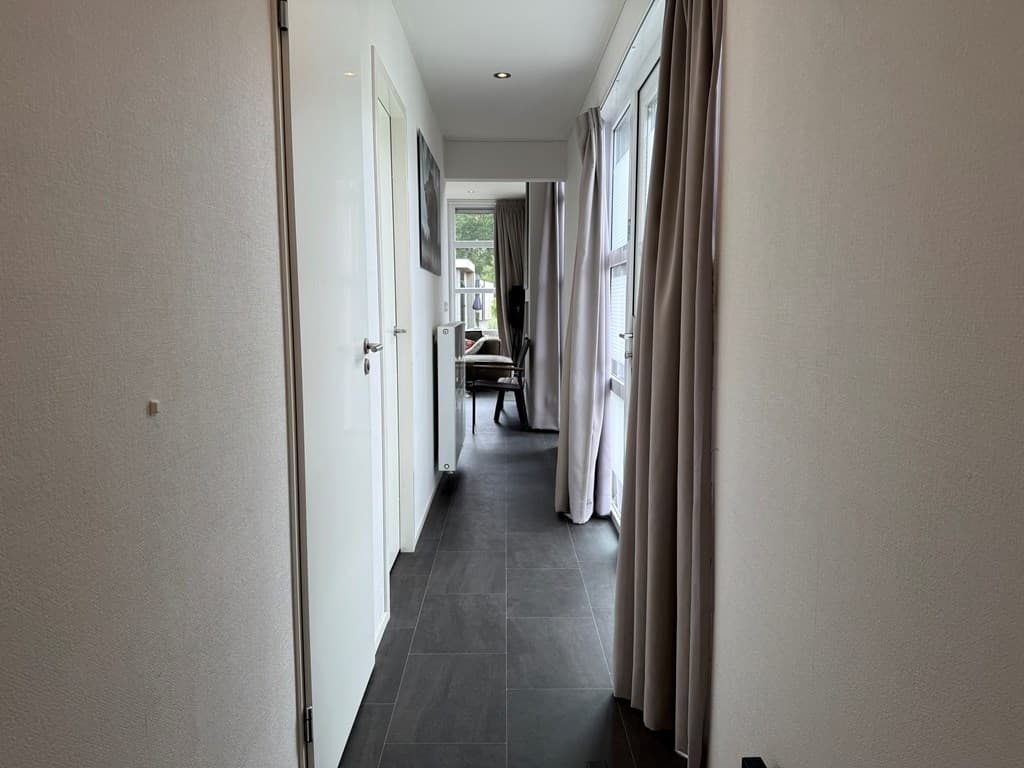 Gang met donkere vloertegels en wanden, voorzien van ramen en gordijnen aan het einde.