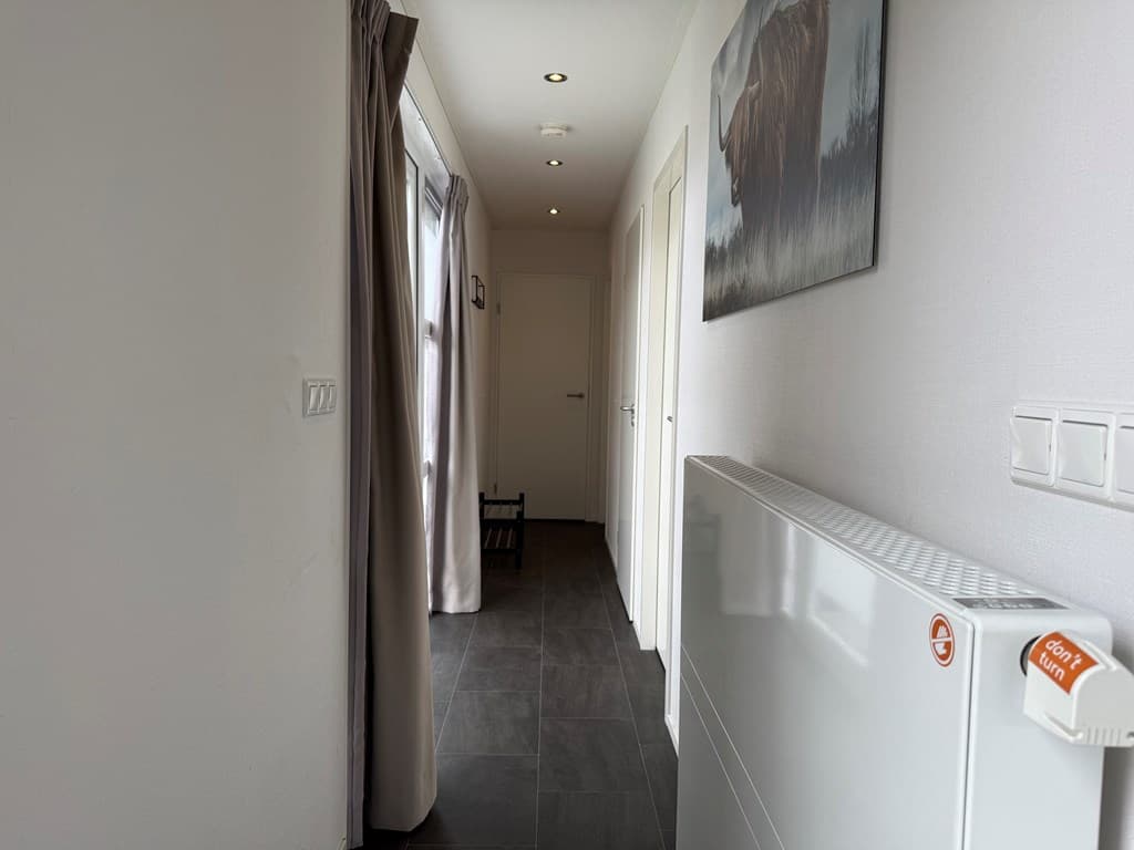 Lichte gang met een donkere tegelvloer, witte muren en een verwarmingsradiator, voorzien van gordijnen en een schilderij.