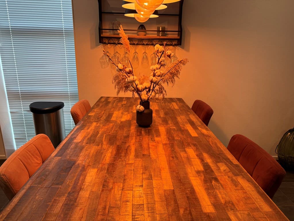 Houten eettafel met zes stoelen en een vaas met gedroogde takken in een lichte ruimte.