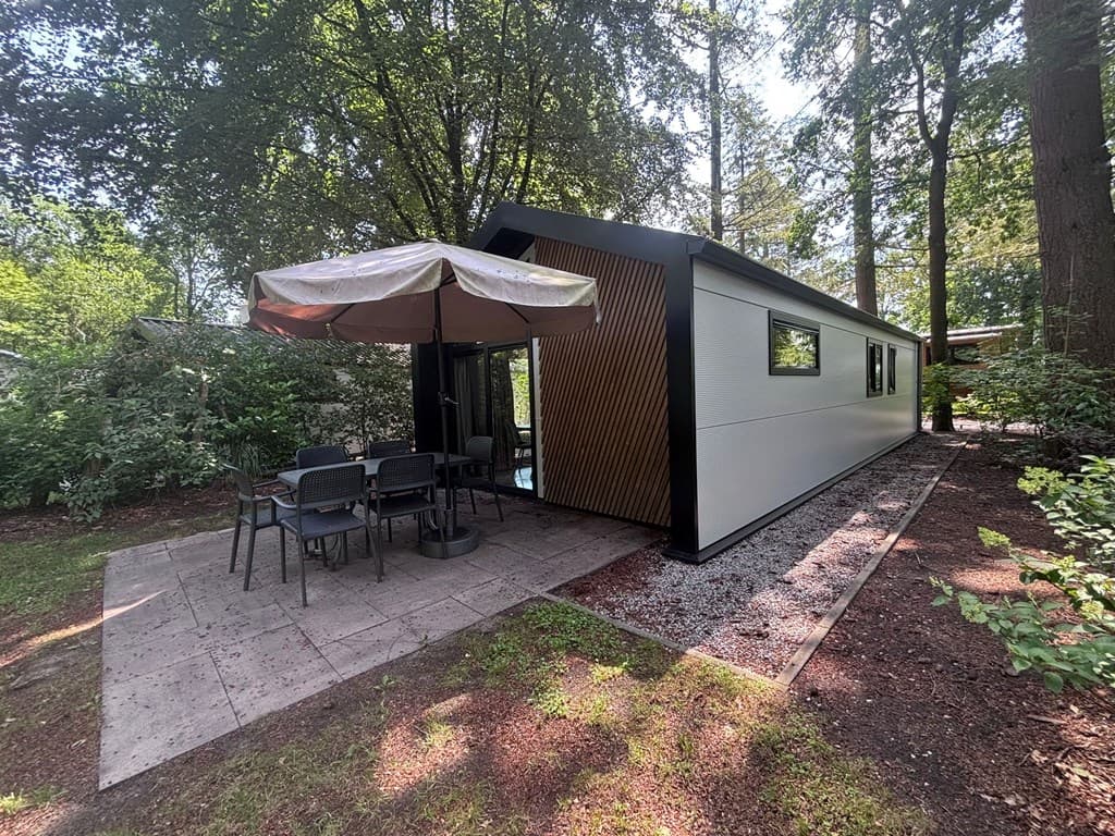 Moderne vakantiewoning met een terras, een grote parasol en een tafel met stoelen, omringd door bomen en groen.