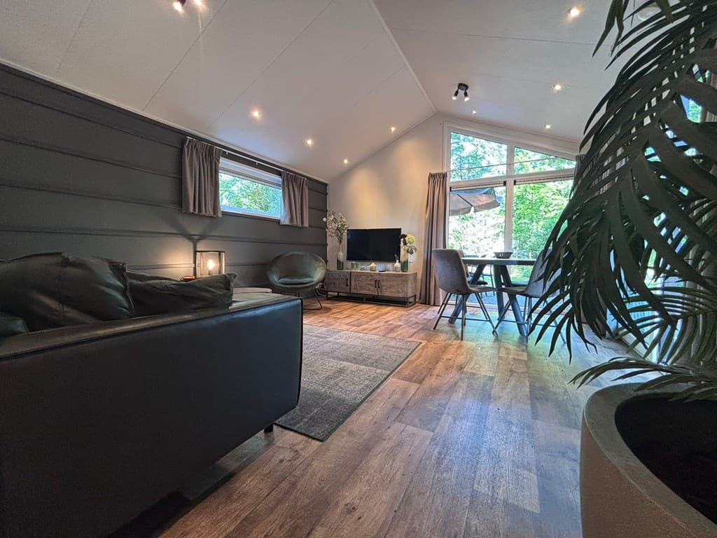 Woonkamer met moderne inrichting, televisie, zithoek, en eettafel met uitzicht op groen door grote ramen