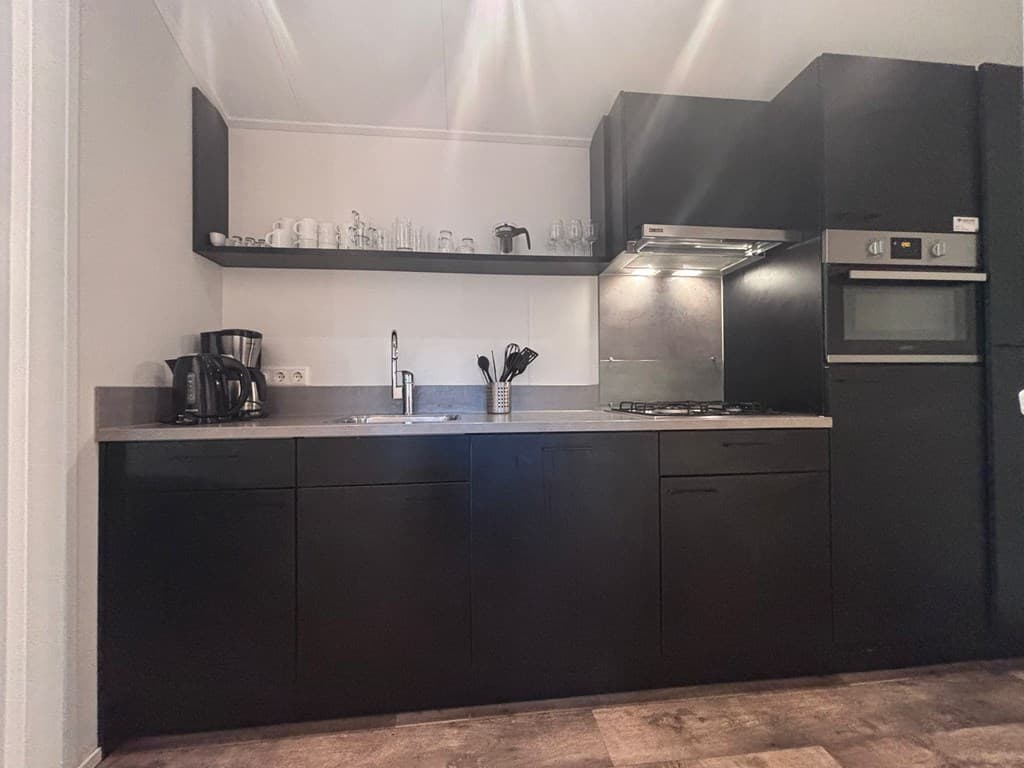 Keuken met zwarte kasten, een spoelbak, kookplaat en bovenkasten met glazen en keukengerei
