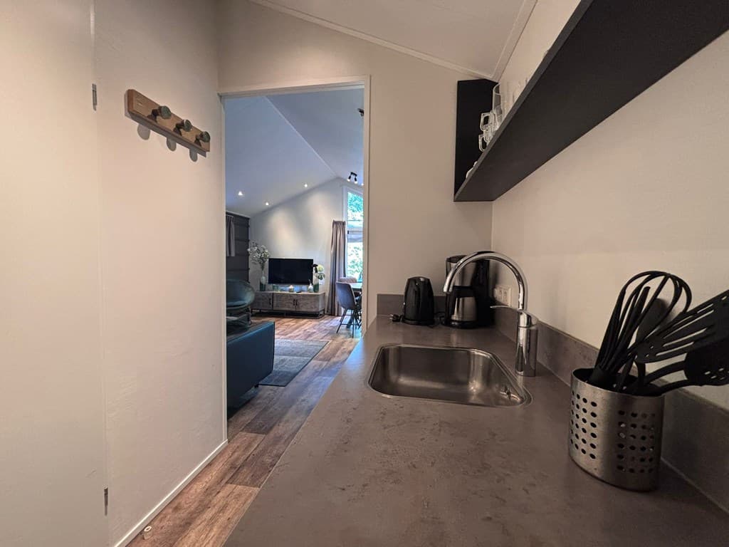 Zicht op een keuken met aanrecht, gootsteen, keukengerei en doorgang naar een woonkamer met televisie en zithoek.