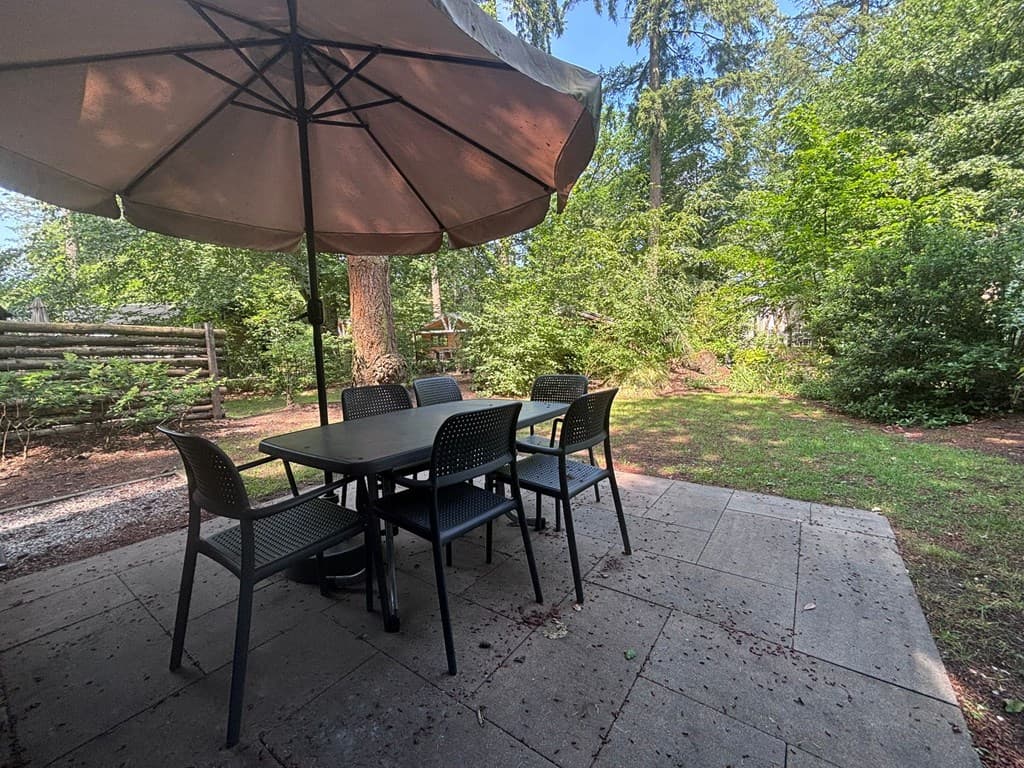 Tuin met een betegeld terras, een tafel met stoelen en een grote parasol, omringd door bomen en groene vegetatie.