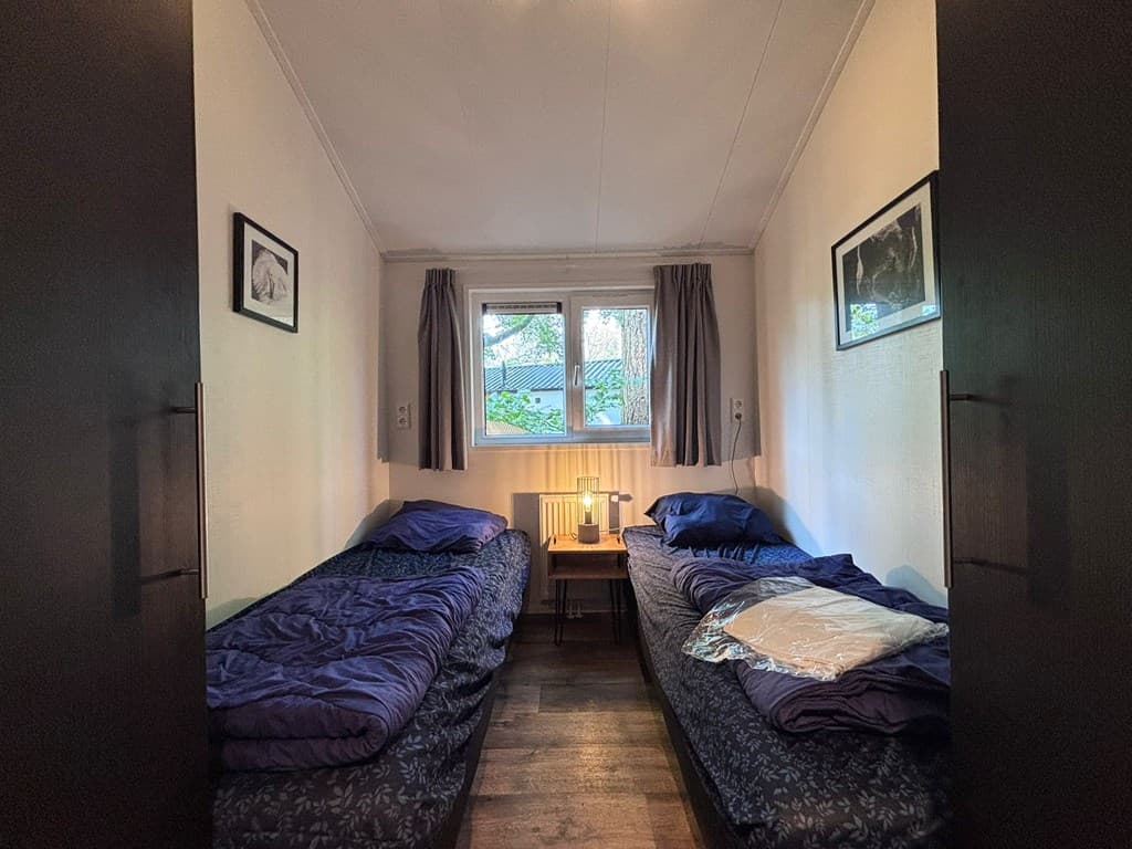 Slaapkamer met twee eenpersoonsbedden, nachtkastje met lamp en raam met uitzicht op bomen.