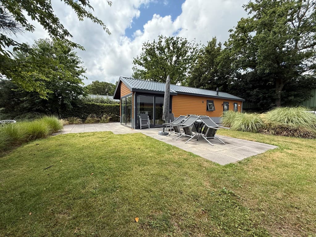 Tuin met een vakantiehuis van EuroParcs Zuiderzee, groen gras, verschillende struiken en een terrasje met ligstoelen.