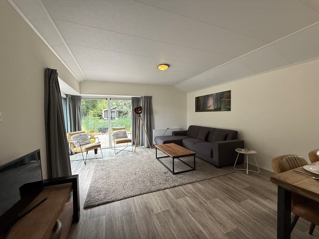 Moderne woonkamer met een bank, salontafel en eetgedeelte, met uitzicht op een terras en groene omgeving buiten.