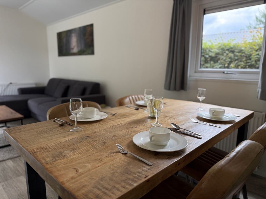 Eettafel met vier borden en glazen, zitbank in de achtergrond en een raam met uitzicht op groen.