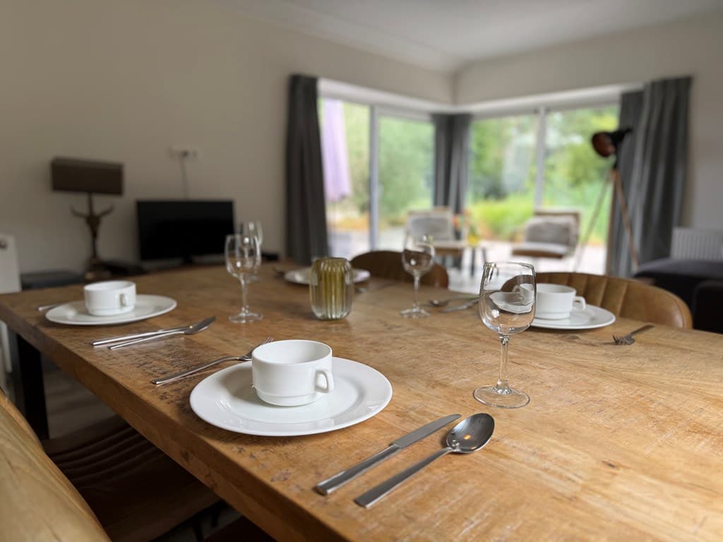 Eettafel met borden, glazen en bestek in een ruime woonkamer met veel natuurlijk licht en uitzicht op de tuin