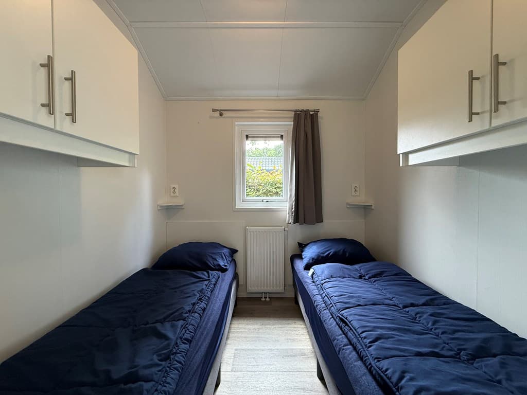 Slaapkamer met twee eenpersoonsbedden, witte kastjes boven de bedden en een raam met gordijnen