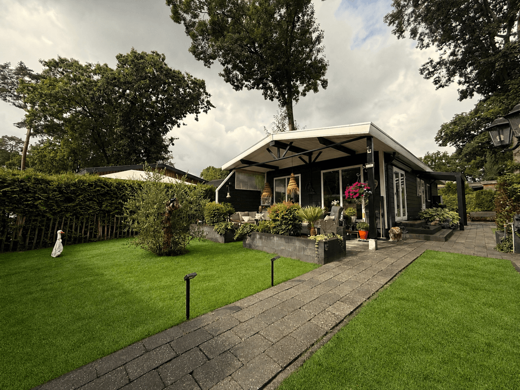 Recreatiewoning met groen gazon, betegeld pad, planten en terrasoverkapping omgeven door bomen en struiken