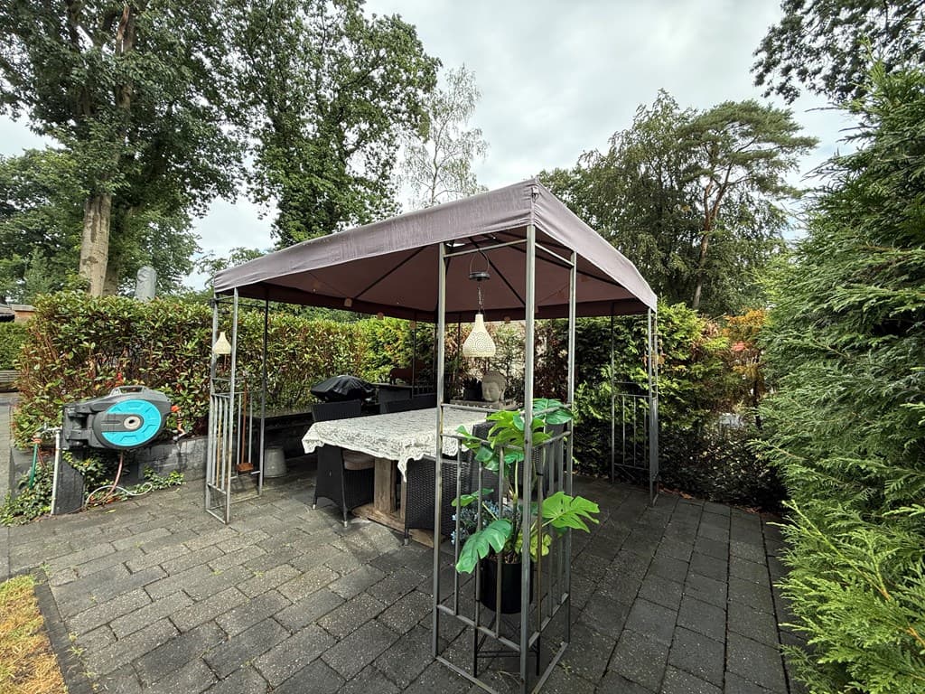 Overdekte buitenruimte met zithoek en tafel, omringd door groene hagen en opvallende planten in een pot.