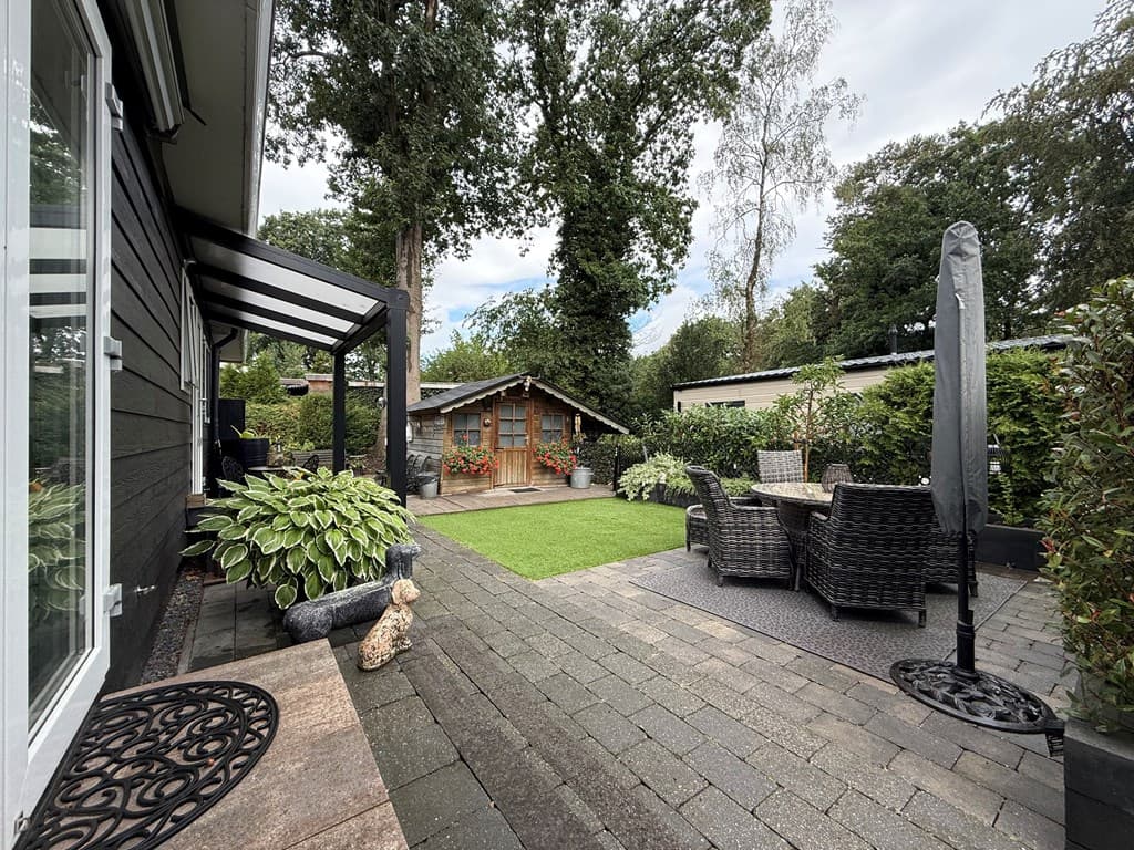 Buitenterrein van de recreatiewoning met terrastegels, tuinmeubilair en een houten tuinschuur omgeven door groen.