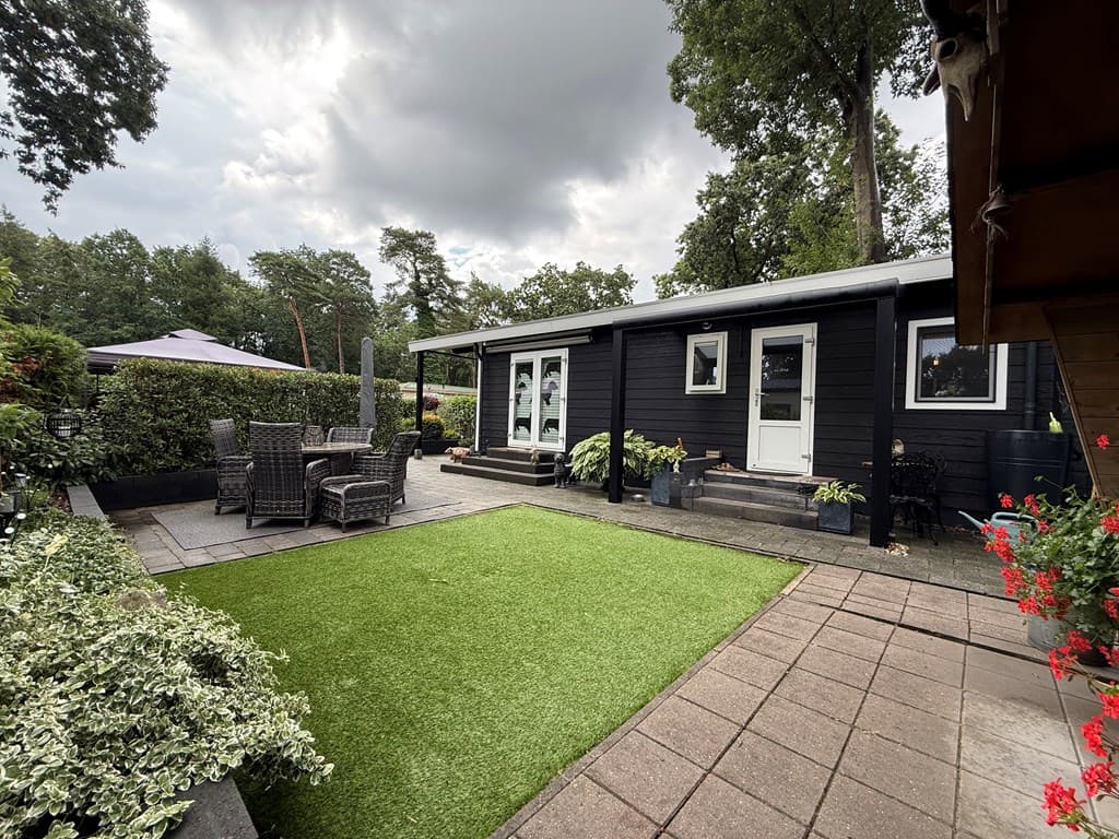 Zwart houten recreatiewoning met terras en groene kunstgrasmat, omringd door bomen en planten.