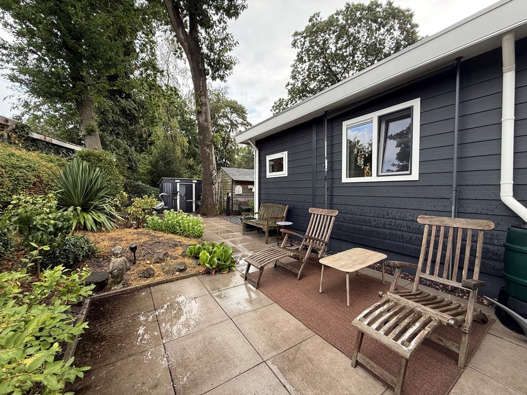 Terras met houten stoelen en tafel, omlijst door groen plantenbed en bomen, aan de achterkant van een bungalow.