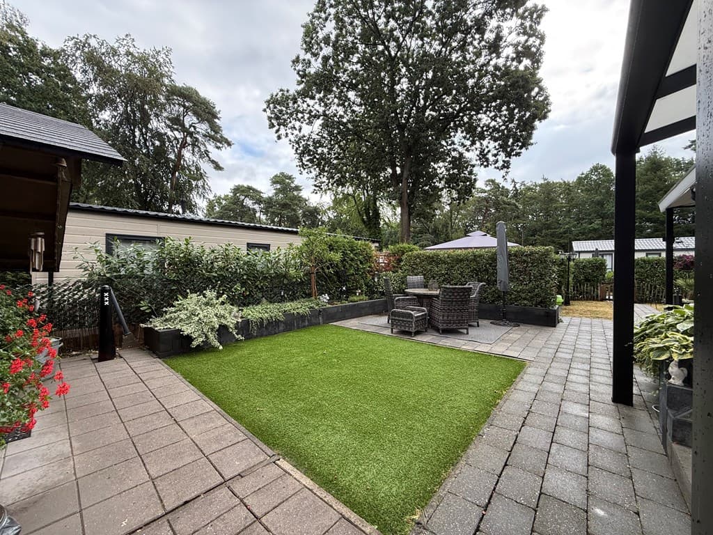Tuin met kunstgras, betegeld pad en zithoek met wicker stoelen, omringd door groene begroeiing en bloemen.