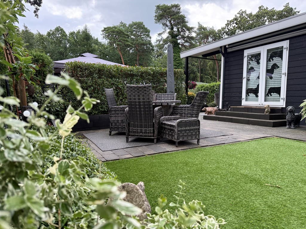 Terras met tuinmeubels van wicker en kunstgras, omringd door struiken en bomen bij een recreatiewoning in Harderwijk