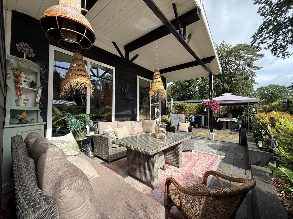 Gedeelte van een terras met een grote loungebank, glazen tafel, hanglampen en planten rondom, en een gazon op de achtergrond