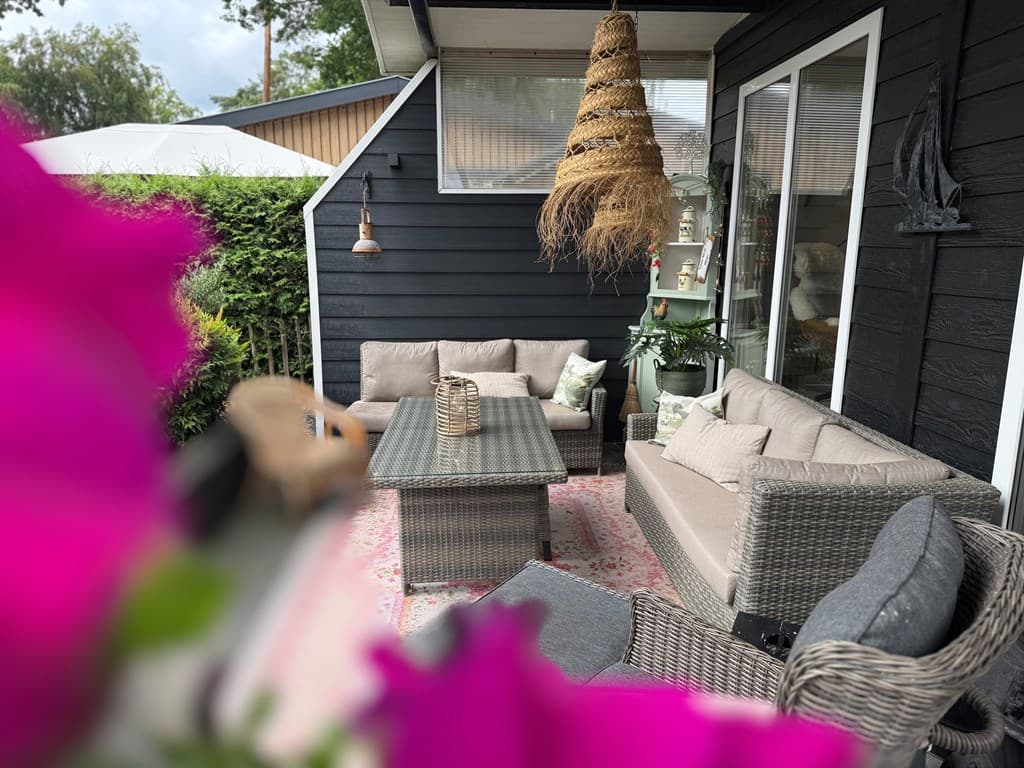 Terras van een recreatiewoning met houten meubels, een rieten hanglamp en groene planten, omgeven door bloemen en struiken.