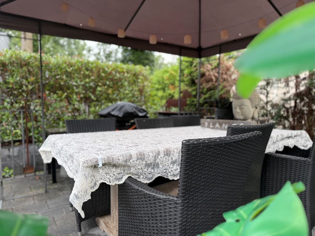 Tuinset met donker wicker meubels onder een pergola, omringd door groene hagen en een decoratieve boeddha.
