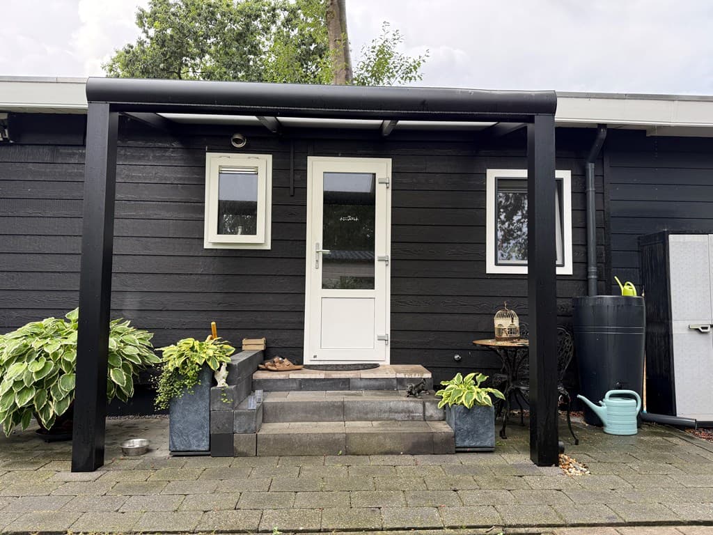 Vooraanzicht van een recreatiewoning met een zwart houten gevel, een witte deur en enkele planten rondom de entree.