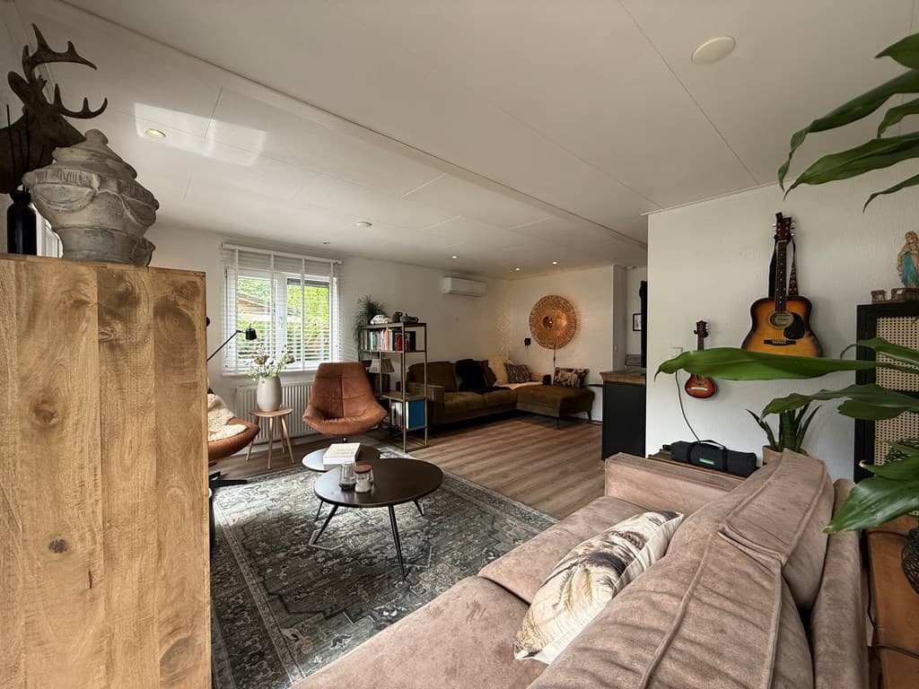 Ruime woonkamer met een zithoek, planten en een ronde tafel, sfeervolle decoraties en verlichting zichtbaar.