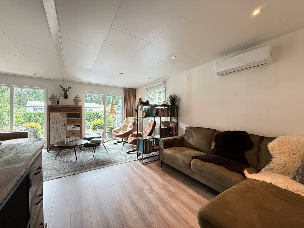 Lichte woonkamer met een bank, boekenkast en fauteuils, grote ramen met zicht op een groene tuin en buitenruimte.
