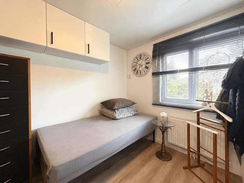 Slaapkamer met een eenpersoonsbed, kastenwand, bureau en raam met jaloezieën in lichte kleuren.