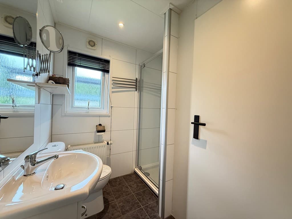 Badkamer met een wastafel, spiegel, douche en toilet, betegelde muren en een raam dat licht binnenlaat