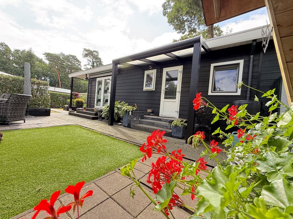 Zwart houten recreatiewoning met terras, bloeiende bloemen en kunstgras in een groene omgeving op Camping De Konijnenberg