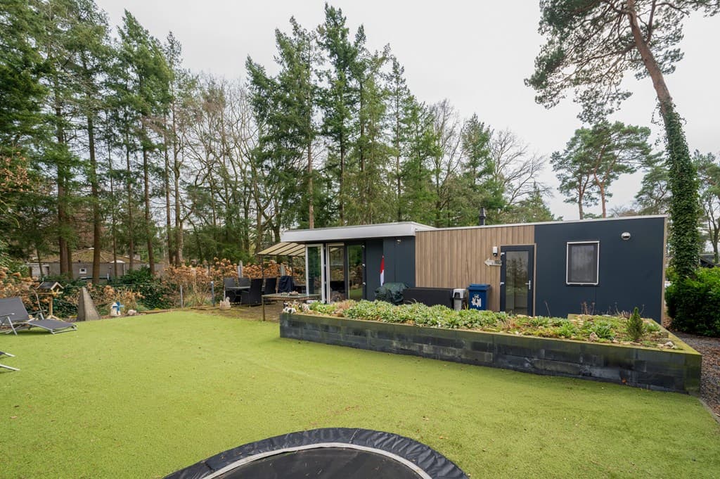 Moderne recreatiewoning met groen grasveld en omringende bomen, inclusief een trampoline en zitgedeelte buiten.