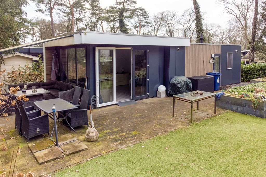 Moderne recreatiewoning met terras, tuinmeubilair en groen gras, omringd door bomen en een schutting.